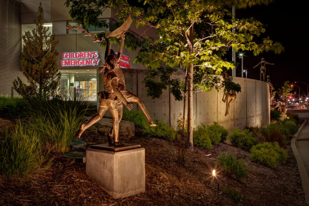 Landscape lighting highlights a bronze statue of children outside a Children's Emergency entrance. 