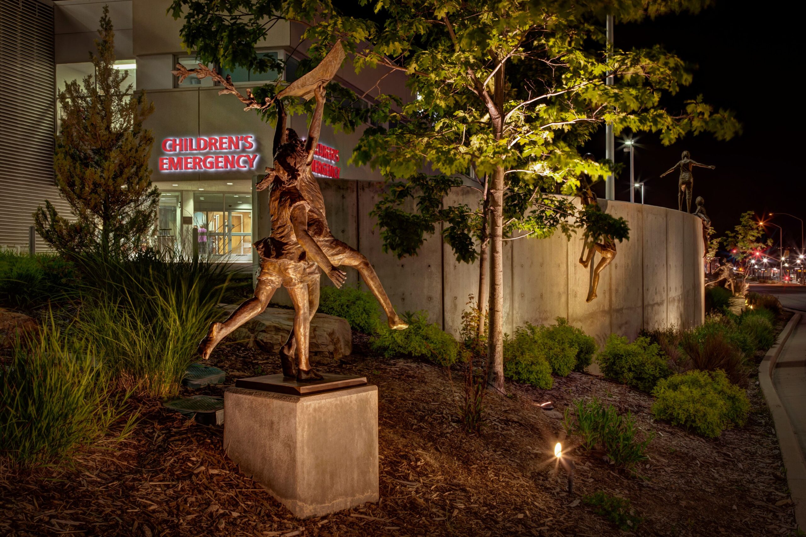 Landscape lighting highlights a bronze statue of children outside a Children's Emergency entrance.