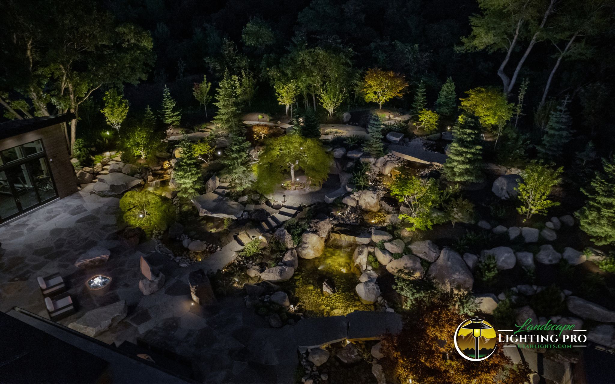 This aerial view of a luxury backyard showcases expertly layered outdoor lighting design. Warm LED lights accentuate stone walkways, lush greenery, and water features, creating a breathtaking nighttime ambiance. Ideal for high-end homes looking to elevate outdoor living after dark.